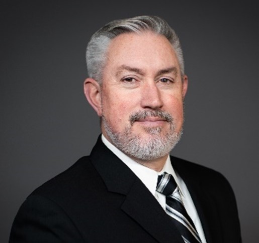 Brent Wallace headshot. He is wearing a dark suit and sits in front of a dark grey background. 