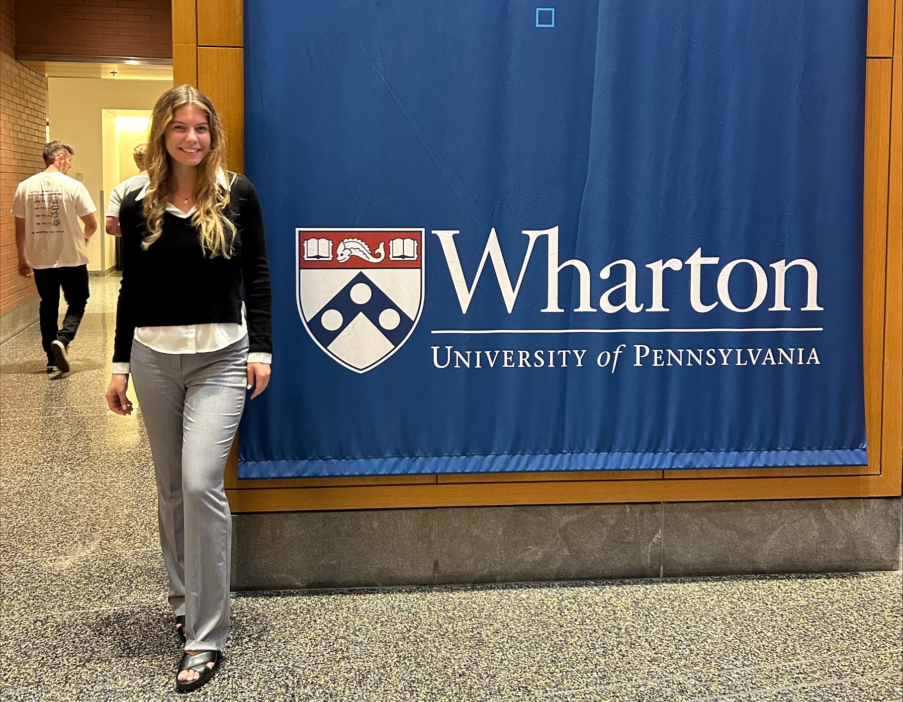 Woman in black shirt and jeans stands next to Wharton College of Busines sign. 